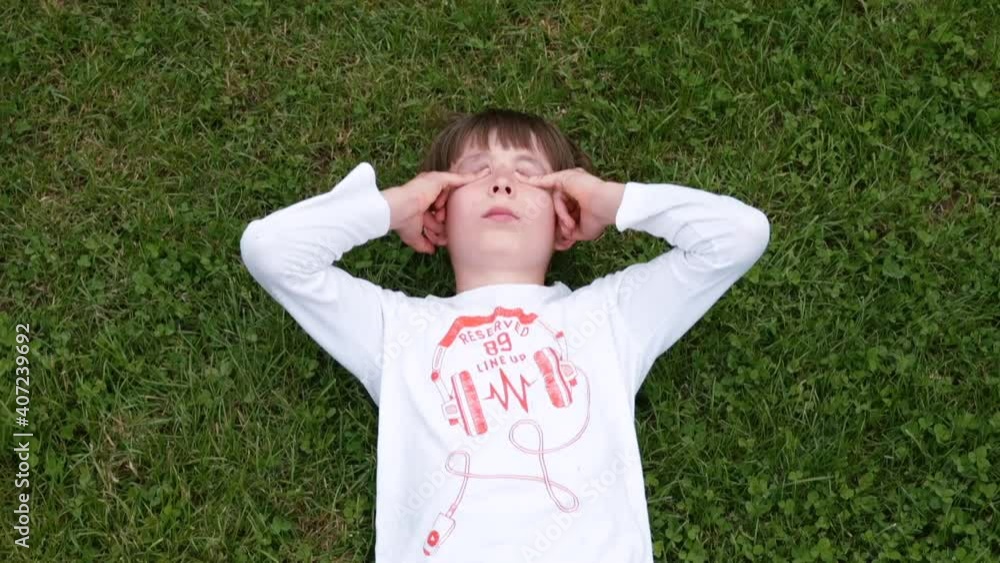 A tired teenager lies on the grass. Rubs eyes with hands, covers ears. The boy's mental experience after the conflict. Relaxation of the psyche. Camera movement. A tired teenager lies on the grass. Rubs eyes with hands, covers ears. The boy's mental experience after the conflict. Relaxation of the psyche. Camera movement.