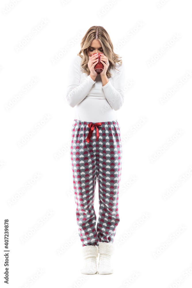 Front view of young woman in pajama drinking coffee from red mug in pajamas after wake up. Full length portrait on white background. Front view of young woman in pajama drinking coffee from red mug in pajamas after wake up. Full length portrait on white background.