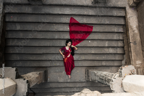 Portrait of beautiful Indian girl in heritage stepwell wearing traditional Indian red saree, gold jewellery and bangles. Maa Durga agomoni shoot concept. Traditional woman on stairs with poses photo