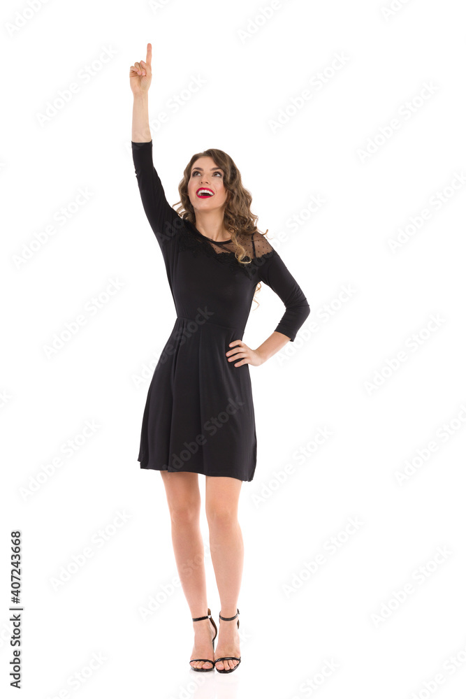Happy Young Woman In Elegant Black Dress And High Heels Is Standing And Pointing Up Happy Young Woman In Elegant Black Dress And High Heels Is Standing And Pointing Up