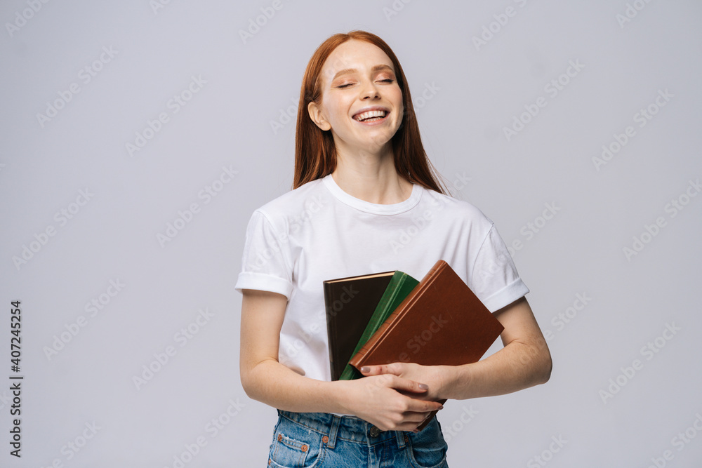 Laughing young woman college student with closed eyes holding books on isolated gray background. Pretty redhead lady model wearing casual clothes emotionally showing facial expressions. Laughing young woman college student with closed eyes holding books on isolated gray background. Pretty redhead lady model wearing casual clothes emotionally showing facial expressions.