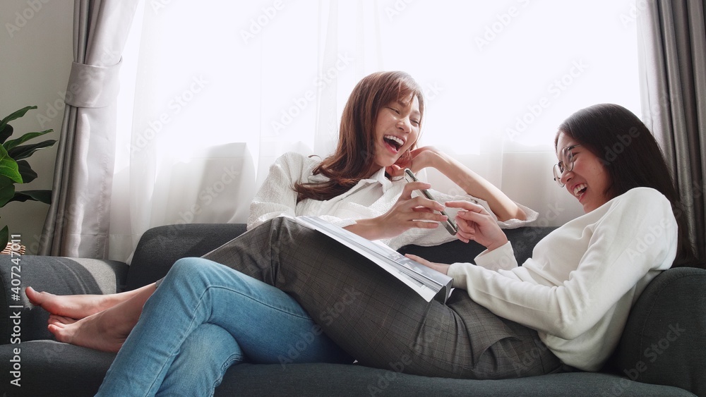 Lovely Asian couple lying on sofa play smartphone with a friend sitting and reading a book together daily holiday life of an Asian woman in the living room at home. Lovely Asian couple lying on sofa play smartphone with a friend sitting and reading a book together daily holiday life of an Asian woman in the living room at home.