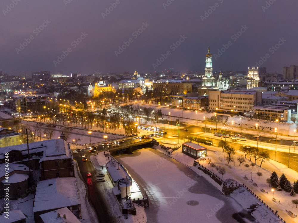 Winter evening illuminated city aerial landscape with frozen snowy river. Lopan embankment, Dormition Cathedral, Serhiivskyi Maidan in Kharkiv, Ukraine