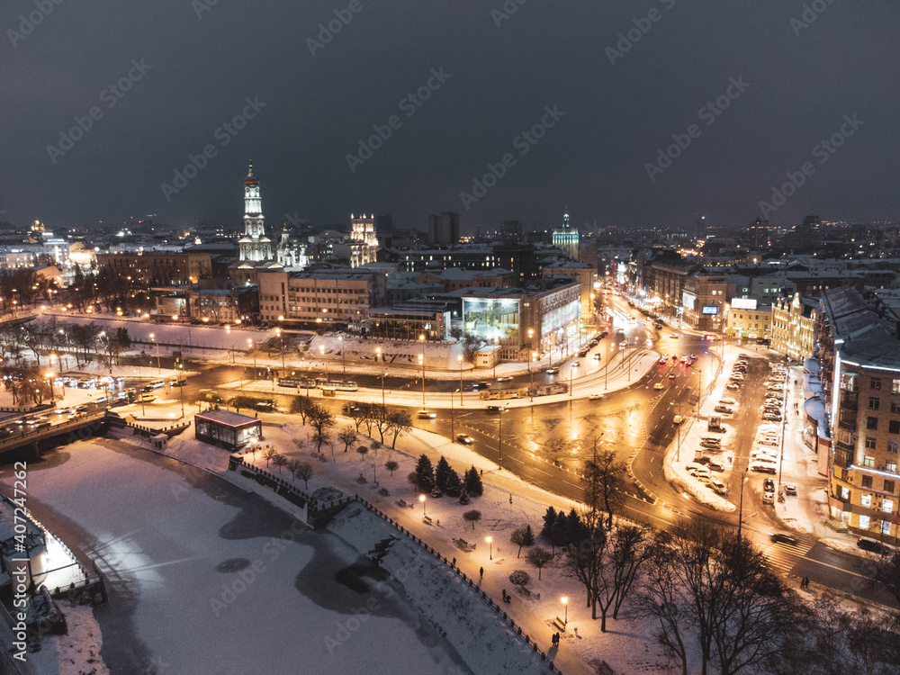 Winter evening illuminated city aerial landscape with frozen snowy river. Lopan embankment, Dormition Cathedral, Serhiivskyi Maidan in Kharkiv, Ukraine