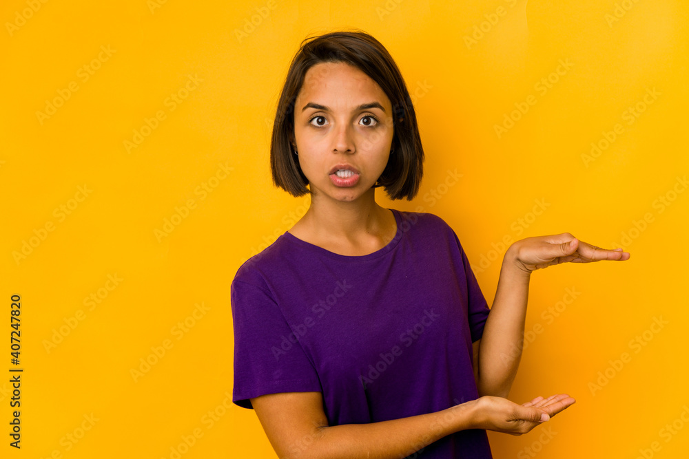 Young hispanic woman isolated on yellow shocked and amazed holding a copy space between hands. Young hispanic woman isolated on yellow shocked and amazed holding a copy space between hands.