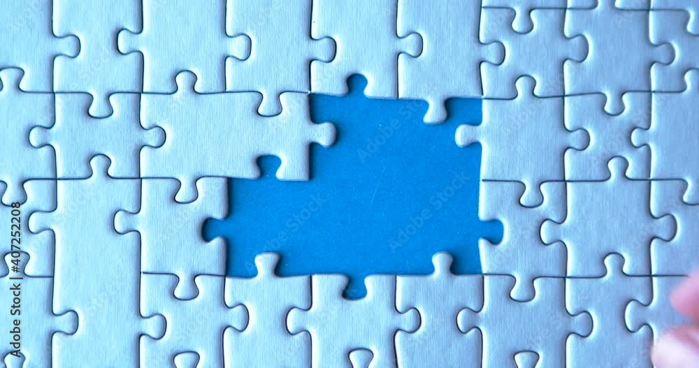 Gray puzzles stacked on blue canvas background. Female hand is putting down pieces. Finishing, completion of work task. Successful achievement of goal. Decision, solution of problem, issue found.