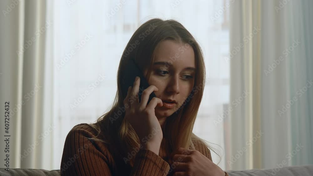 Young girl in brown sweater and blue jeans sits on sofa in front of the window and annoyed talking on a mobile phone. Young girl in brown sweater and blue jeans sits on sofa in front of the window and annoyed talking on a mobile phone.