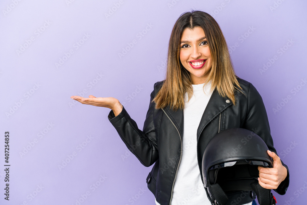Young indian motorbiker woman isolated showing a copy space on a palm and holding another hand on waist. Young indian motorbiker woman isolated showing a copy space on a palm and holding another hand on waist.