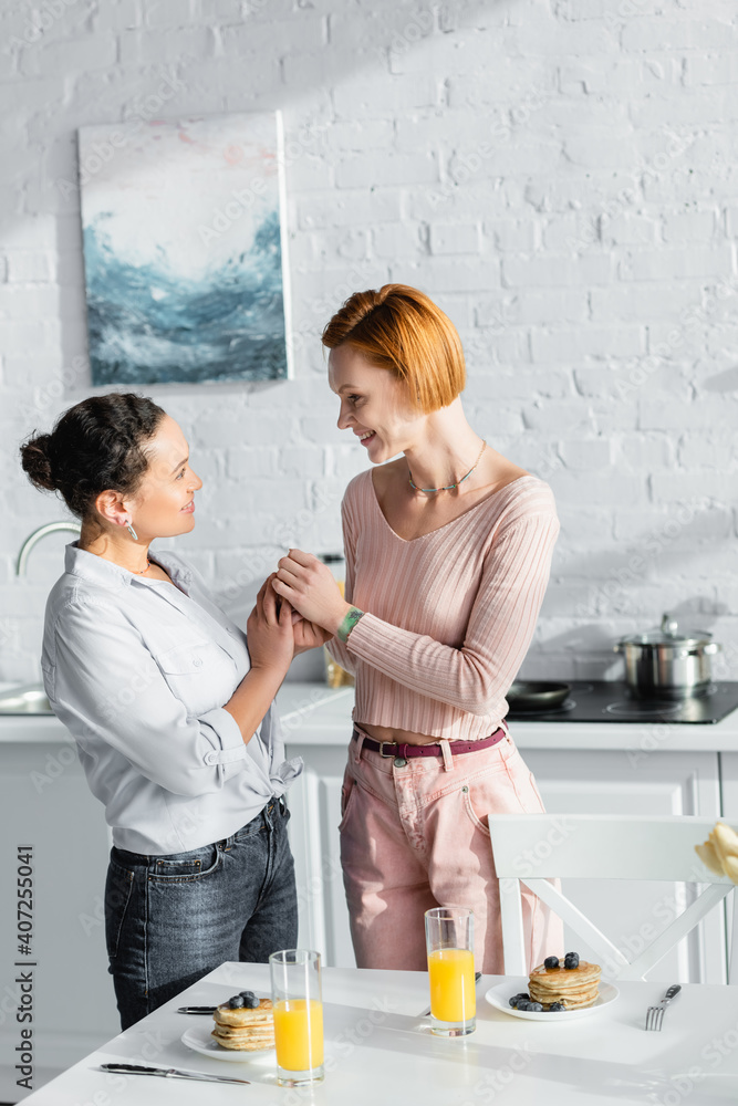 smiling interracial lesbian couple holding hands and looking at each other near breakfast on table