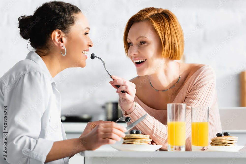 cheerful lesbian woman feeding african american girlfriend with blueberry during breakfast cheerful lesbian woman feeding african american girlfriend with blueberry during breakfast