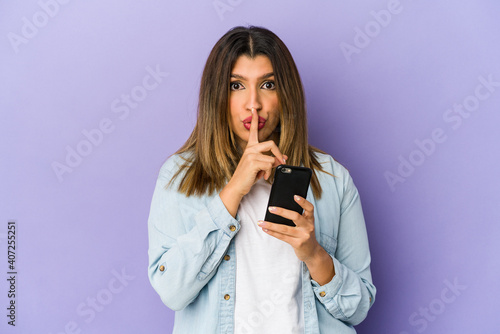Young indian woman holding a phone isolated keeping a secret or asking for silence.