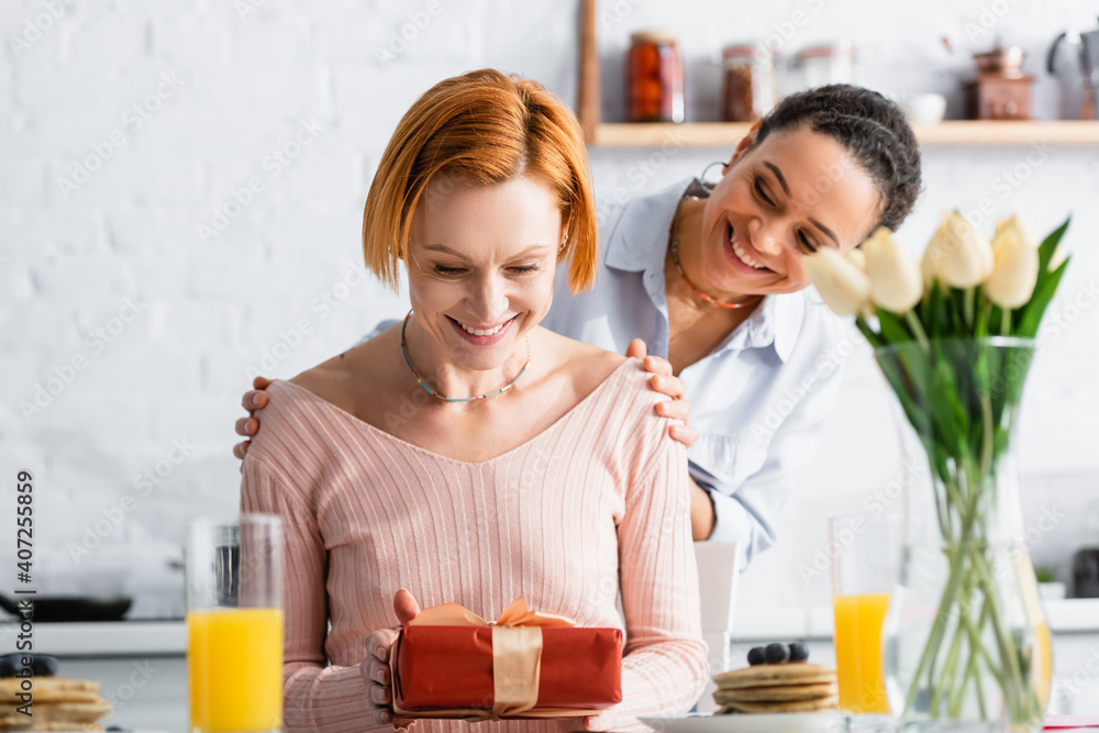 smiling african woman hugging shoulders of happy lesbian girlfriend holding gift on valentines day, blurred foreground smiling african woman hugging shoulders of happy lesbian girlfriend holding gift on valentines day, blurred foreground