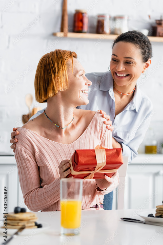 smiling african american woman embracing shoulders of happy lesbian girlfriend holding present near breakfast on blurred foreground smiling african american woman embracing shoulders of happy lesbian girlfriend holding present near breakfast on blurred foreground