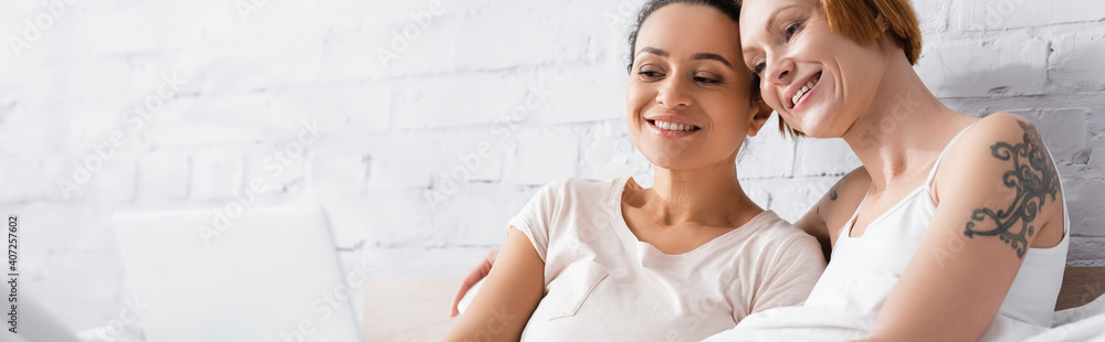 cheerful interracial lesbian couple watching movie on computer in bed, banner cheerful interracial lesbian couple watching movie on computer in bed, banner
