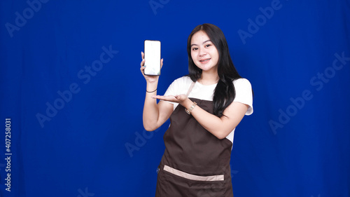 asian woman shopkeeper pointing blank screen handphone isolated blue background