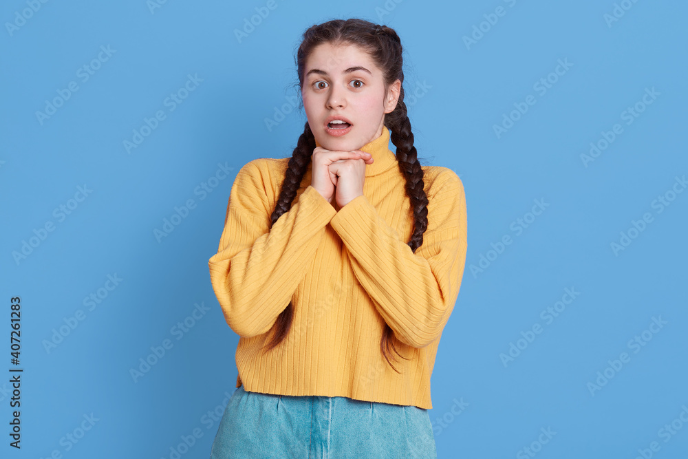 Young beautiful brunette woman wearing yellow sweater, looks shocked, keeping fists under chin and mouth opened, standing isolated over blue background. Young beautiful brunette woman wearing yellow sweater, looks shocked, keeping fists under chin and mouth opened, standing isolated over blue background.