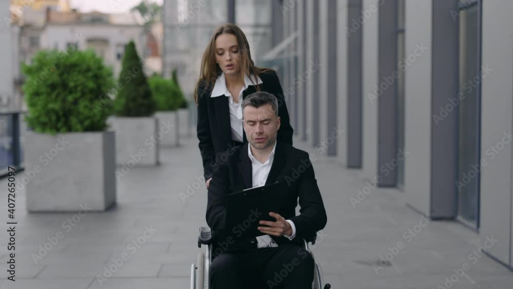 Crop view of young woman pushing wheelchair with disabled boss and stopping near office building. Male businessman looking and showing documents to female coworker while talking. Crop view of young woman pushing wheelchair with disabled boss and stopping near office building. Male businessman looking and showing documents to female coworker while talking.