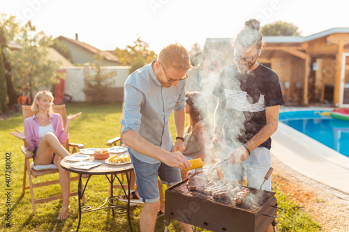 Canvas Print Men grilling meat while women are  sitting and relaxing by the pool at backyard