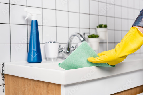 Cropped view of woman in rubber glove cleaning bathroom sink with rag near bottle of detergent on blurred background