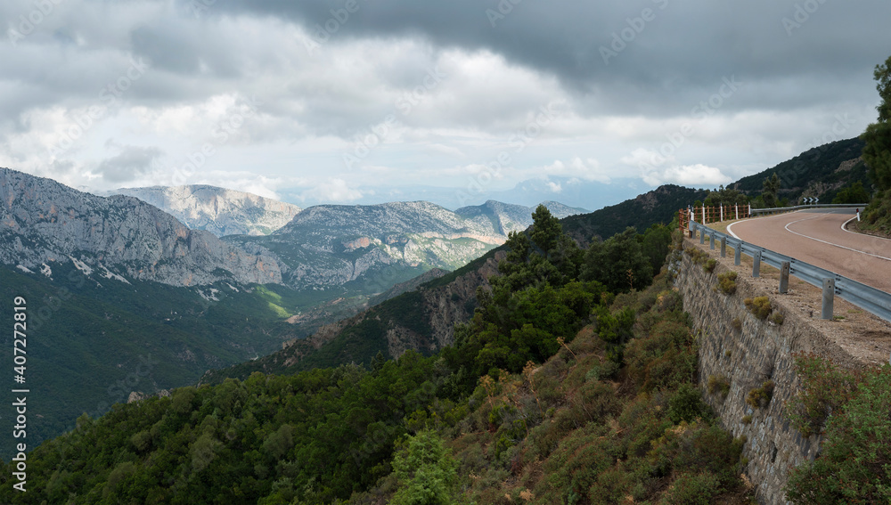 Panoramic view of landscape of Supramonte Mountains with winding asphalt road, green hills, trees and mediterranean forest vegetation. Ogliastra, Sardinia, Italy. Summer cloudy sky