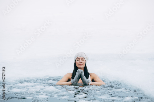 Winter swimming. Woman in frozen lake ice hole. Swimmers wellness in icy water. How to swim in cold water. Beautiful young female in zen meditation. Gray hat and gloves swimming clothes. Nature lake © Girts