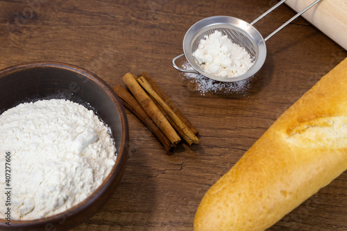 Flour in a wooden bowl, small sieve, French baguette and cinnamon sticks on a wooden table. Side view with copy space. Kitchen baking tools with place for text, or logo.