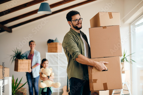 Canvas Print Happy man carrying stack of carton boxes while moving with his family into new home