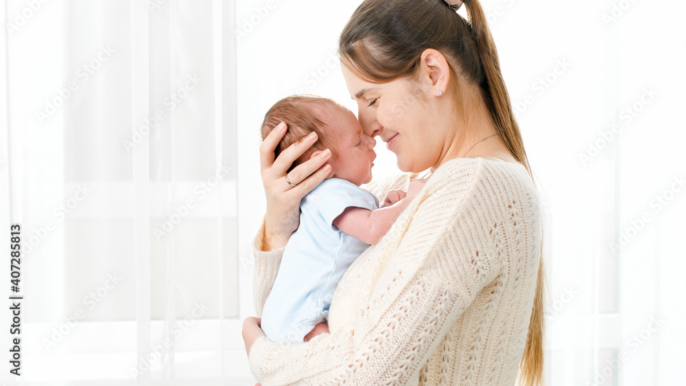 Portrait of happy smiling mother cuddling and looking on her newborn baby son against big window at house. Concept of family happiness and loving parents with little children. Portrait of happy smiling mother cuddling and looking on her newborn baby son against big window at house. Concept of family happiness and loving parents with little children.