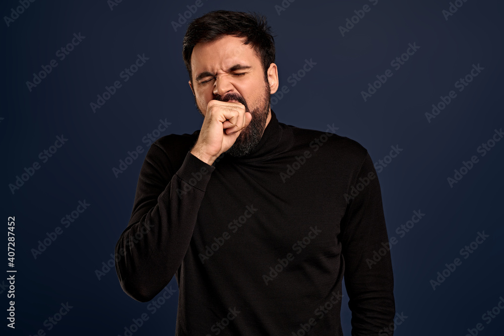 Handsome guy being bored talking with stranger about random staff, yawning cover opened mouth with fist, squinting tired, standing with fatigue look, dont like boring events, blue background