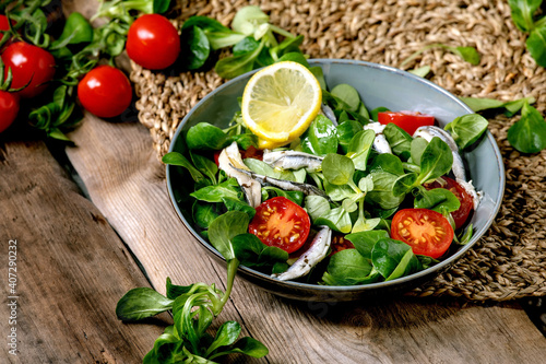 Green field salad with pickled anchovies or sardines fillet, and cherry tomatoes, served in blue bowl with lemon and olive oil on straw napkin over old wooden table.