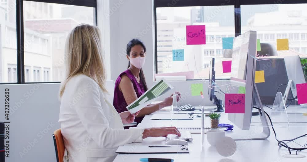 Diverse businesswomen wearing face masks using computers passing paperwork in office Diverse businesswomen wearing face masks using computers passing paperwork in office