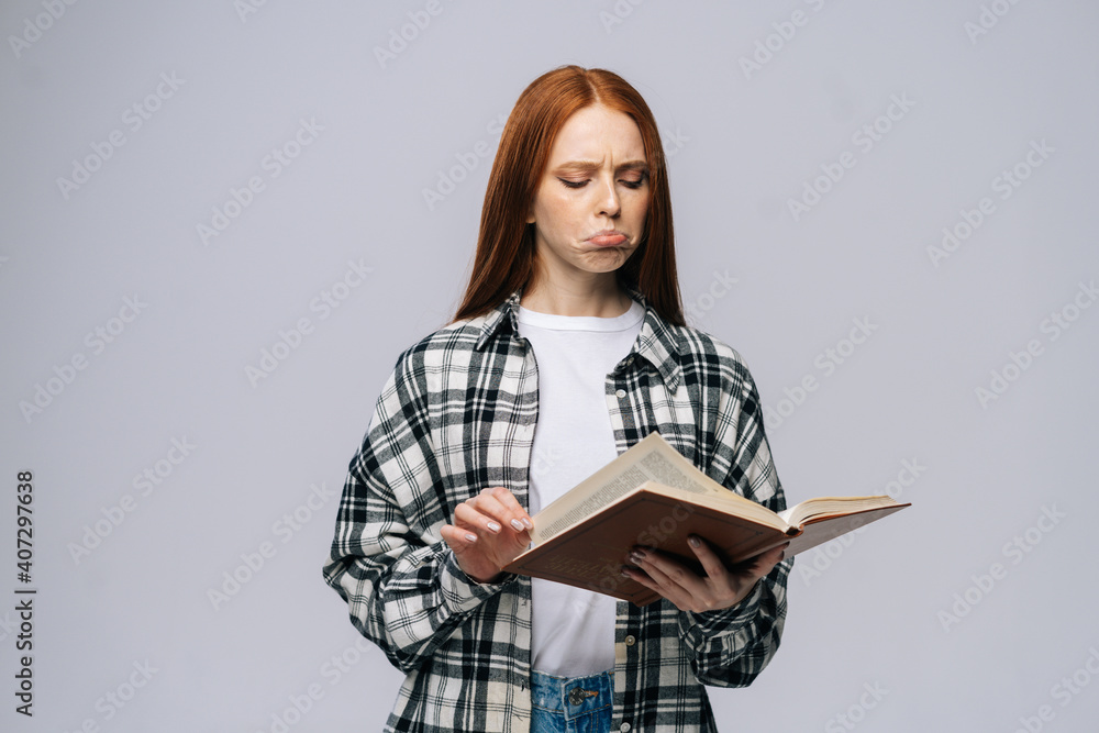 Obraz premium Confused young woman college student turning pages of book on gray isolated background. Pretty redhead lady model wearing casual clothes emotionally showing facial expressions in studio, copyspace.