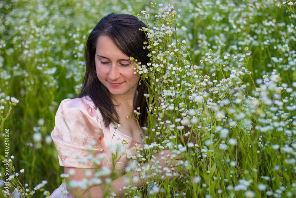 Portrait of a beautiful happy girl with dark hair in pink dress in white wildflowers in the fild at golden time. Young smiling woman sitting on the grass and among the many small flowers. Portrait of a beautiful happy girl with dark hair in pink dress in white wildflowers in the fild at golden time. Young smiling woman sitting on the grass and among the many small flowers.