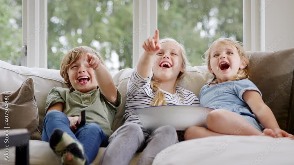 Three children sitting on sofa at home laughing and watching TV with popcorn together - shot in slow motion Three children sitting on sofa at home laughing and watching TV with popcorn together - shot in slow motion