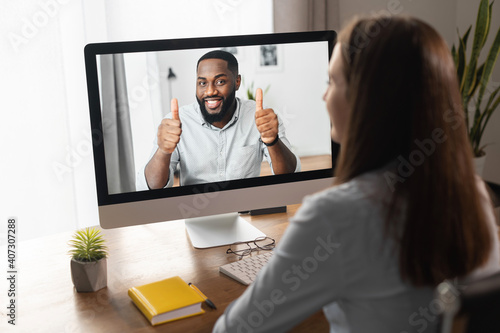 An African-American online coach  mentor  tutor on the laptop screen shows thumbs up. Back view a woman watching webinar  has video meeting with a biracial man
