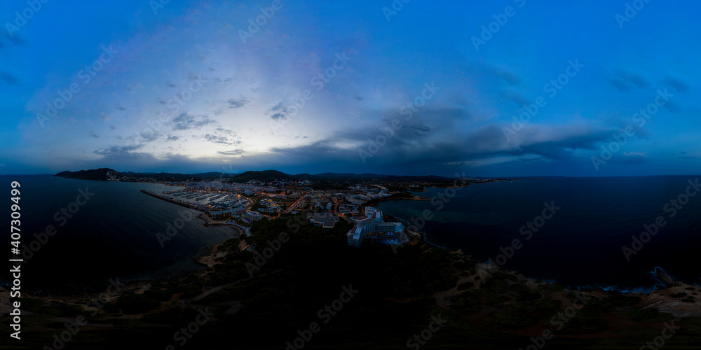 Aerial drone 360 Degree panoramic sphere aerial photo of the beautiful beach front of Ibiza in Spain showing the Spanish beach and hotels from above in the evening with the sun setting over the hills
