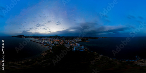 Aerial drone 360 Degree panoramic sphere aerial photo of the beautiful beach front of Ibiza in Spain showing the Spanish beach and hotels from above in the evening with the sun setting over the hills