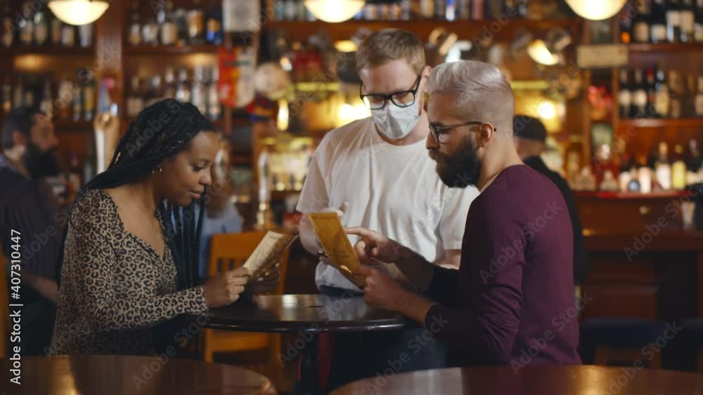 Waiter in safety mask taking order from romantic diverse couple sitting at bar table Waiter in safety mask taking order from romantic diverse couple sitting at bar table