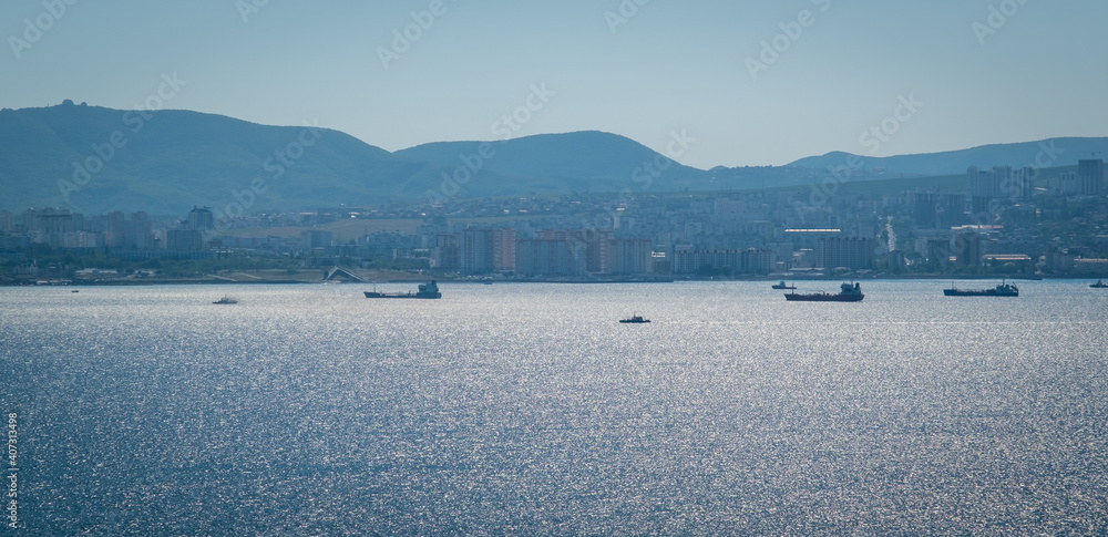 View of Novorossiysk and the port of Novorossiysk. Summer landscape on a sunny day. The sun is reflected in the water. Port cranes in the background