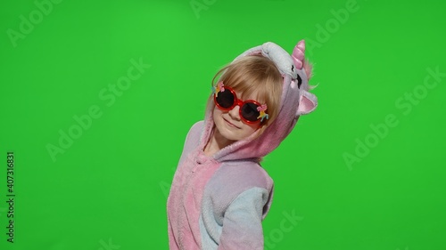 Portrait of a cute young little child in sunglasses smiling, dancing in unicorn costume on chroma key green background. Kid girl animator making gun gesture with hands in unicorn pajamas. Copy space
