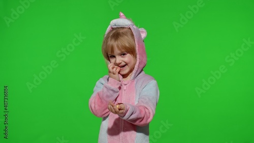 Portrait of a cute little child smiling  enjoying eating sweets candies dessert in unicorn costume on chroma key green background. Model kid girl animator in unicorn pajamas. Copy space