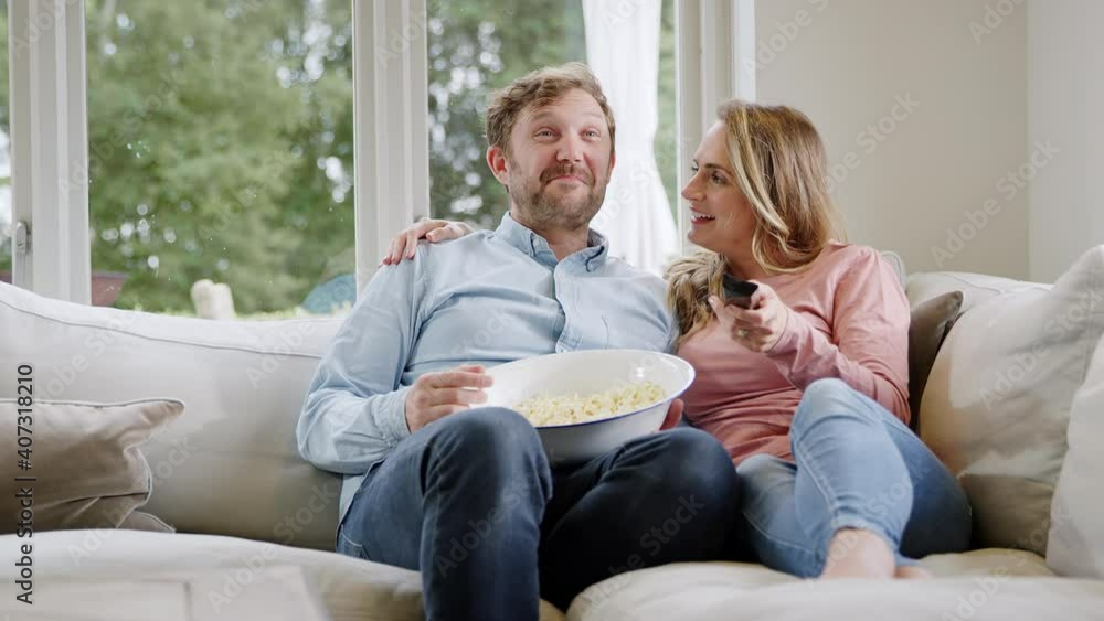 Couple on date night sitting on sofa at home laughing and watching movie on TV with popcorn together - shot in slow motion Couple on date night sitting on sofa at home laughing and watching movie on TV with popcorn together - shot in slow motion