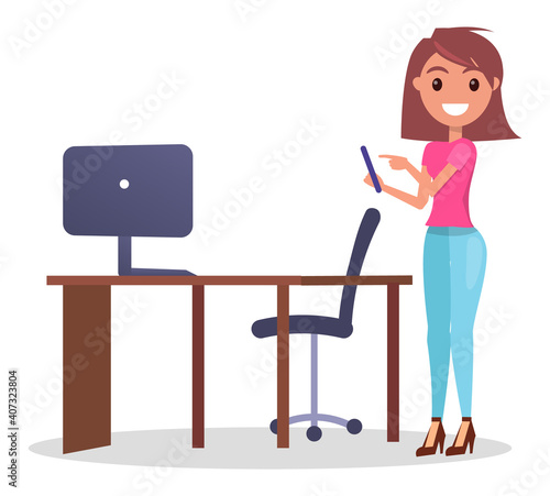 Young pretty girl using phone standing near office table with computer and armchair. Smiling woman point show at screen of phone. Cheerful woman wearing pink t-shirt and jeans using mobile phone
