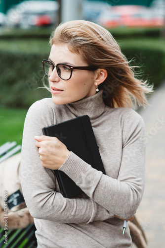 Soft focus student with glasses sits on a bench, reads books and writes notes. A girl with blue eyes, soft skin and long blond hair dreams of a luxurious lifestyle on a fresh spring background.