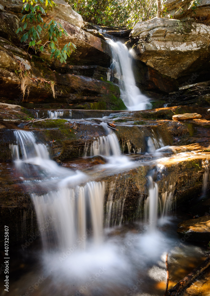 Fototapeta premium Small waterfall cascades over sunlit rocks in summer forest
