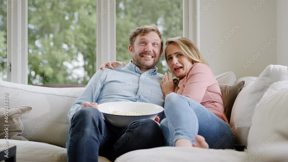 Couple on date night sitting on sofa at home and watching scary movie on TV with popcorn together - shot in slow motion Couple on date night sitting on sofa at home and watching scary movie on TV with popcorn together - shot in slow motion