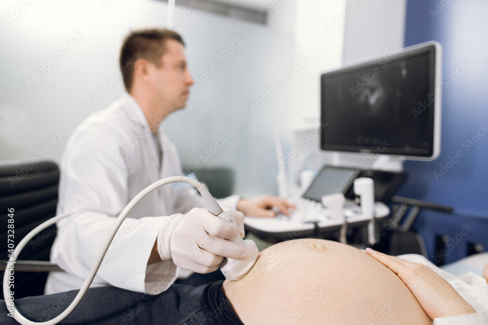 Close-up shot of male doctor performing Ultrasound Sonogram procedure to unrecognizable pregnant woman. Focus on the ultrasound transducer on the belly of the future mother