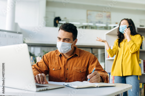 Successful male manager or student wearing protective medical mask, is working on a business project using a laptop, in the background a colleague talks on the phone in a protective mask