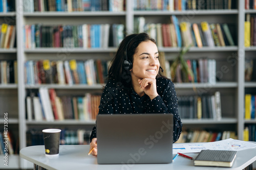 Pretty formal caucasian young woman, call center worker, student, or successful business tutor, in a headset is using a laptop while working online sitting at her work desk and looks away and smiling