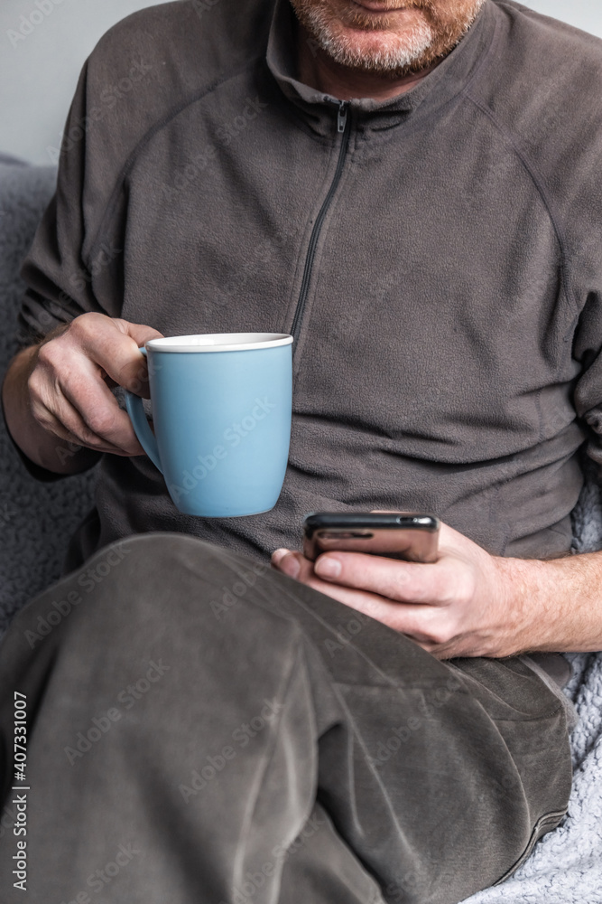 A man sits on the couch at home and drinks tea, texting with friends and relatives using a mobile phone. Close-up. A man sits on the couch at home and drinks tea, texting with friends and relatives using a mobile phone. Close-up.